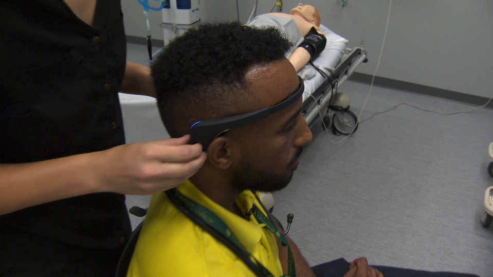 Patient seated in ER simulation as clinician adjusts a headband brain-monitor; gurney with mannequin visible in background