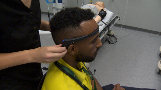 Patient seated in ER simulation as clinician adjusts a headband brain-monitor; gurney with mannequin visible in background