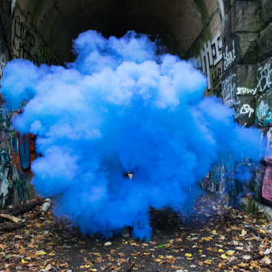 Blue smoke cloud fills a graffiti-covered stone tunnel, partly obscuring a person holding a smoke canister.
