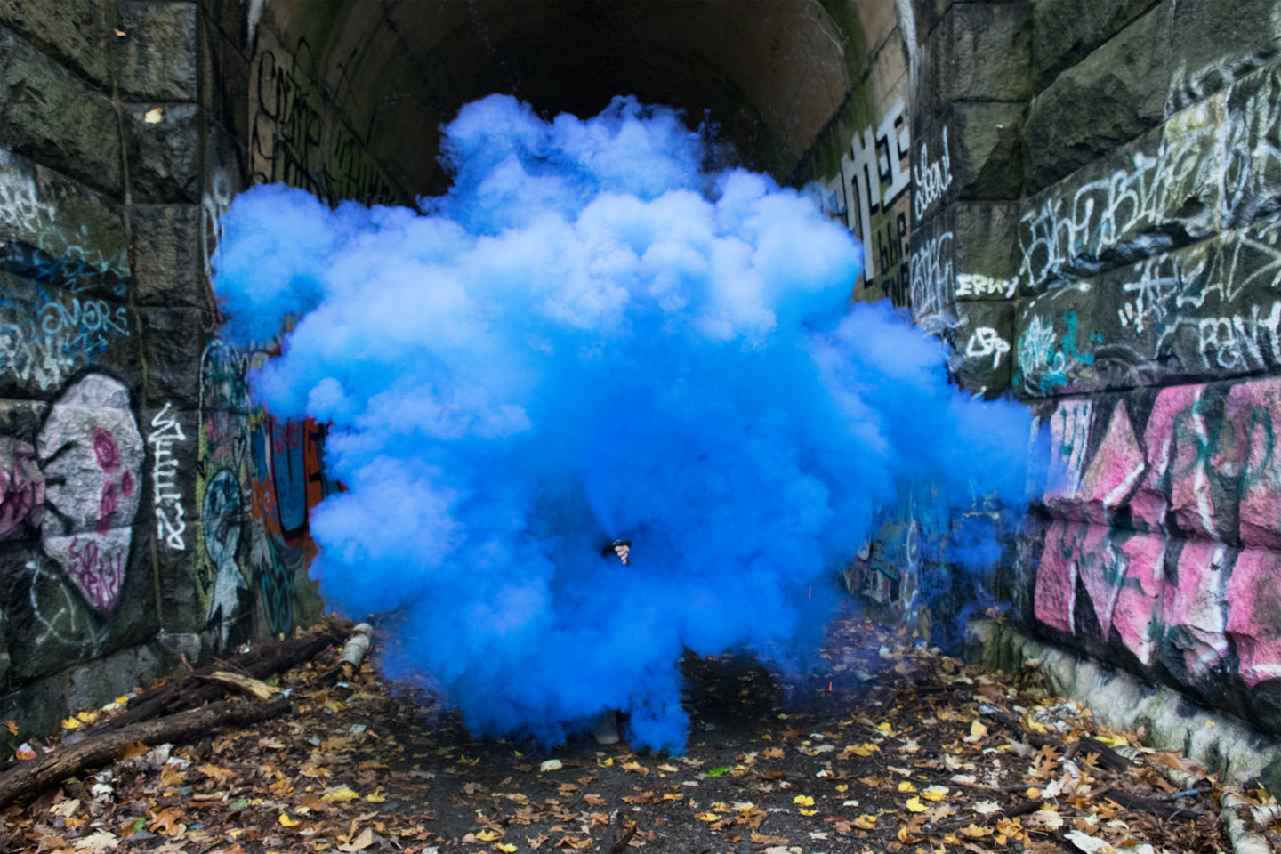 Blue smoke cloud fills a graffiti-covered stone tunnel, partly obscuring a person holding a smoke canister.