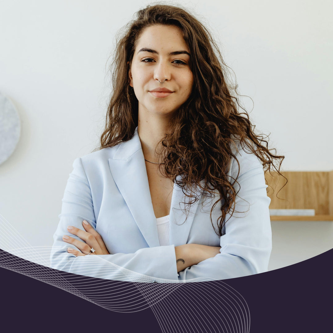 Confident young woman with long curly hair in a light-blue blazer, arms crossed, office background.