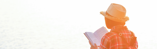 Person wearing straw hat and backpack reading a book by a sunlit lakeshore