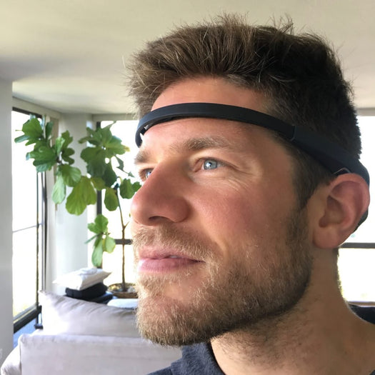 Man wearing a black EEG headband, looking toward window in a living room with a fiddle-leaf fig plant