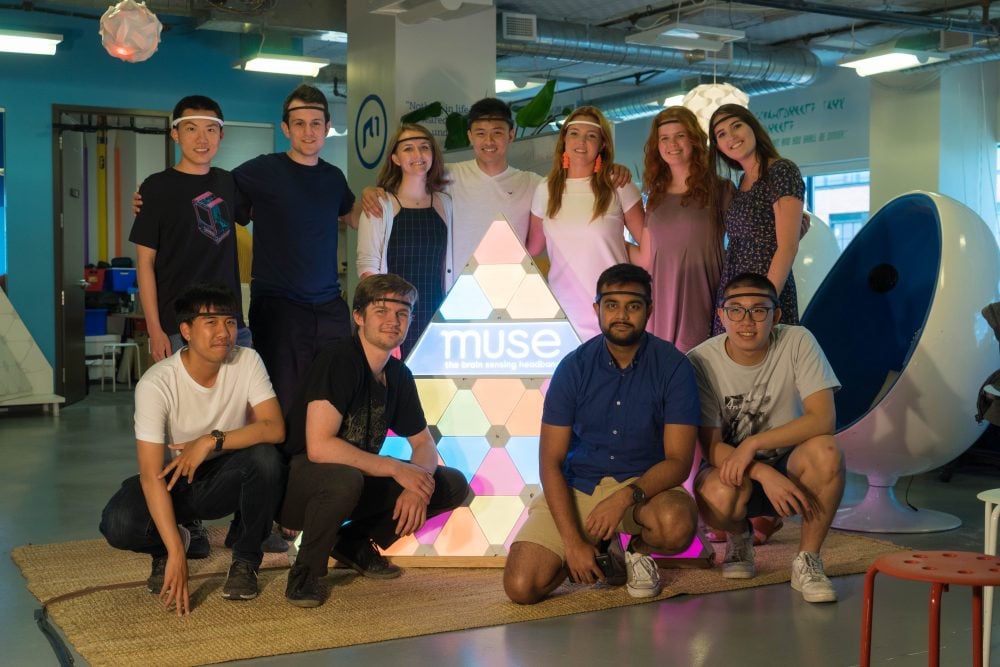 Group of Muse interns posing around an illuminated triangular display reading muse the brain sensing headband in an office setting