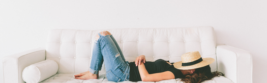 Woman lying on a white couch with a straw sunhat covering her face, wearing jeans and a black top