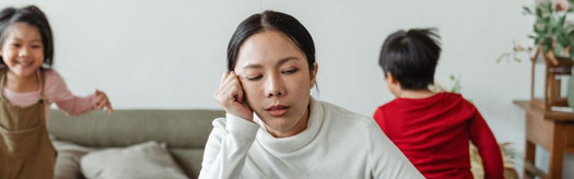 Tired mother resting her head while two children play on a living room couch behind her