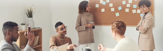 Five coworkers collaborating at a meeting table, discussing ideas by a corkboard of sticky notes.
