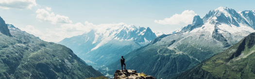 Person standing on a rocky outcrop overlooking snow-capped mountains and a deep valley