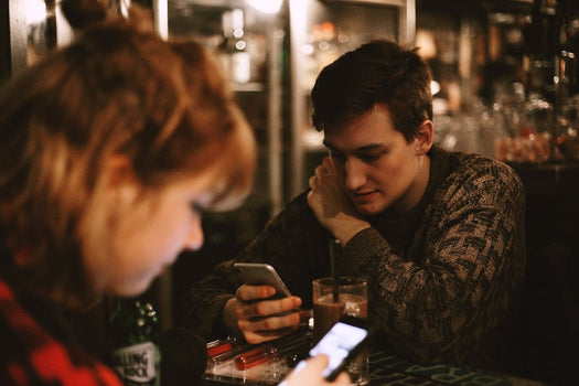 Two young adults at a dim café table, both looking down at their smartphones.