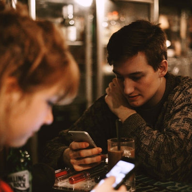 Two young adults at a dim café table, both looking down at their smartphones.