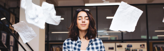 Young woman with eyes closed meditating as papers float around her in a library-like workspace