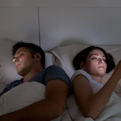 Couple lying back-to-back in bed each looking at their phone, illuminated by screen light