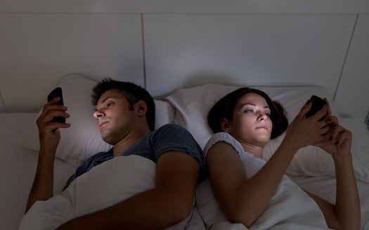 Couple lying back-to-back in bed each looking at their phone, illuminated by screen light