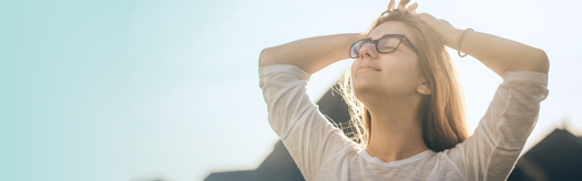 Young woman with glasses outdoors, eyes closed and arms raised, practicing mindfulness in sunlight