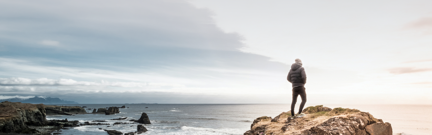 Person standing on rocky coastline looking toward the sea at sunrise with distant cliffs and a cloudy sky