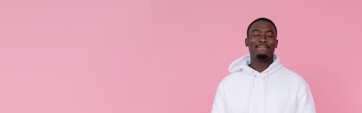 Relaxed young Black man in white hoodie with eyes closed, meditating against a pink background.