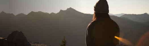 Person wearing a beanie seated on a ridge, looking toward sunlit mountain range at dusk