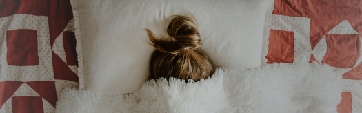 Top of a person's head with a messy bun peeking from under a white duvet, red-and-white geometric quilt at edges.