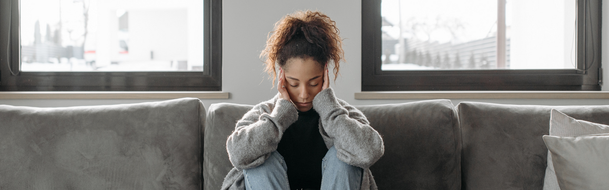 Woman seated on a couch holding her head in her hands, appearing stressed in a bright living room