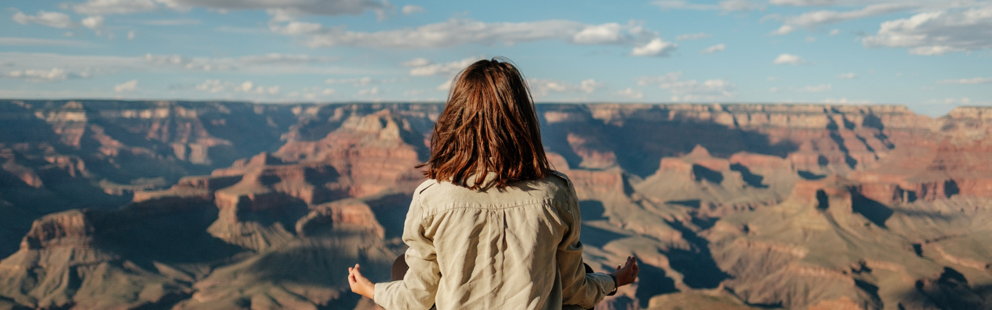 Person seated cross-legged, meditating on a canyon rim with expansive canyon and blue sky.