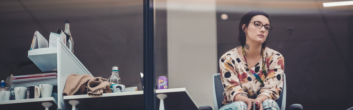 Young woman sitting in an office meeting room looking disengaged, hands clasped, personal items and a purple Twist can behind glass