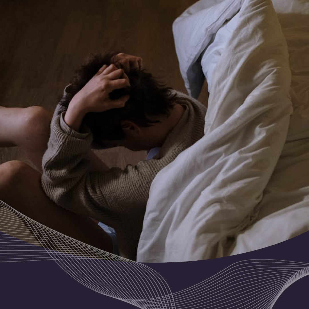 Person sitting on a bed holding their head in their hands in a dim bedroom, appearing anxious
