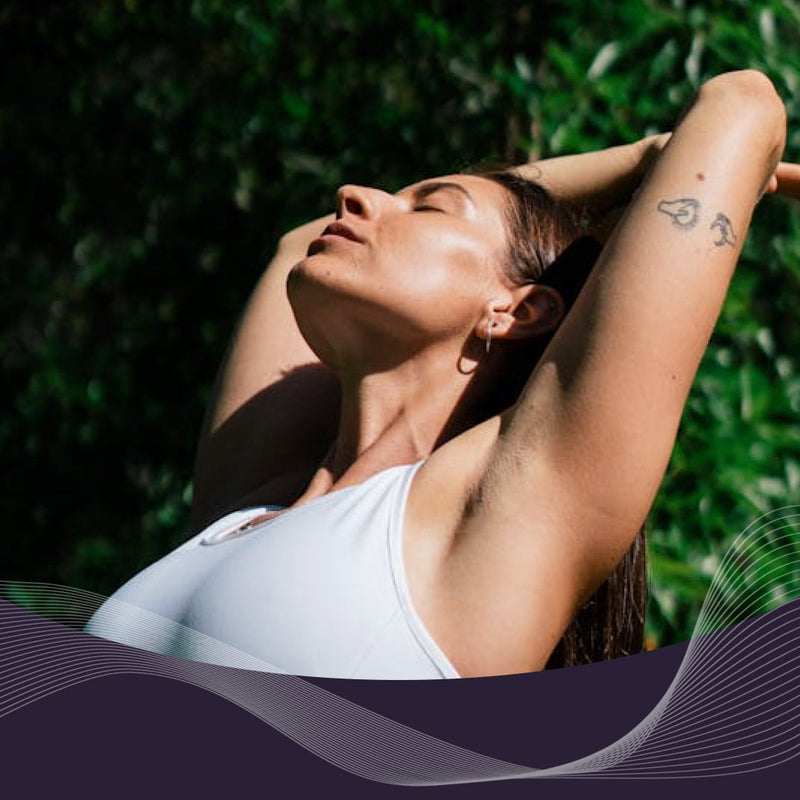 Woman in white tank top stretching with arms raised and eyes closed in a sunlit leafy garden, appearing relaxed