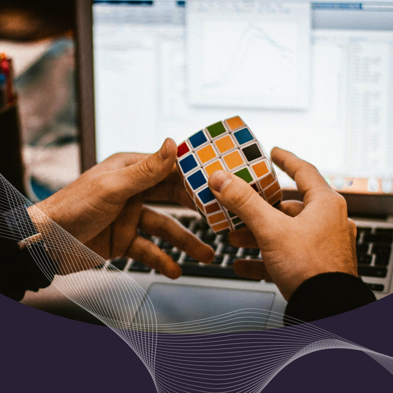 Hands twisting a multi-colored Rubik-style puzzle in front of a laptop with a faint chart and a purple wave overlay at the bottom