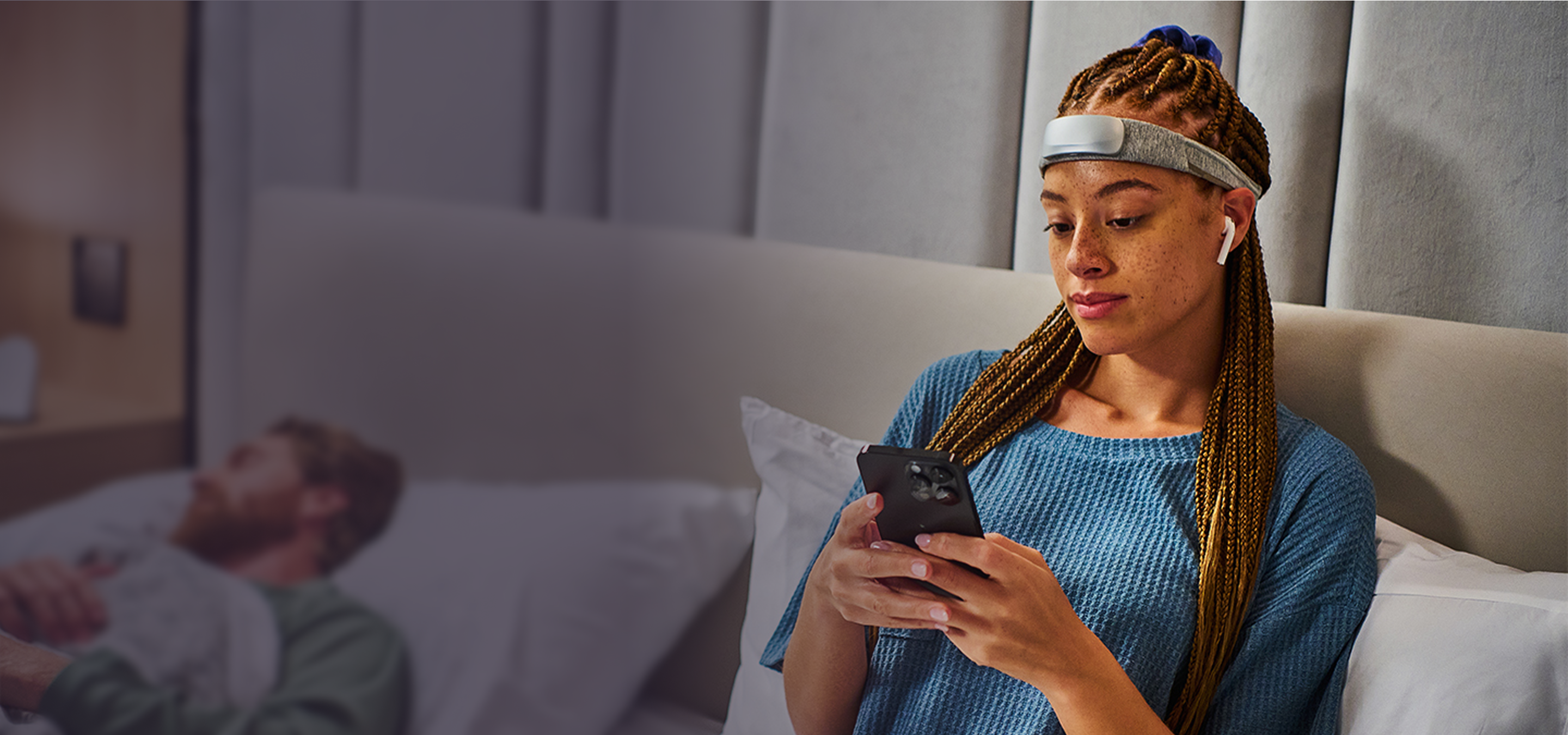 Woman with braided hair wearing a headband device and AirPods, using a smartphone on a bed; man sleeps blurred behind her.