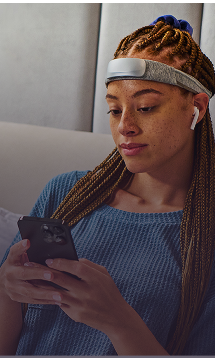 Woman wearing a headband-style forehead wearable and wireless earbud, looking at her smartphone while seated.