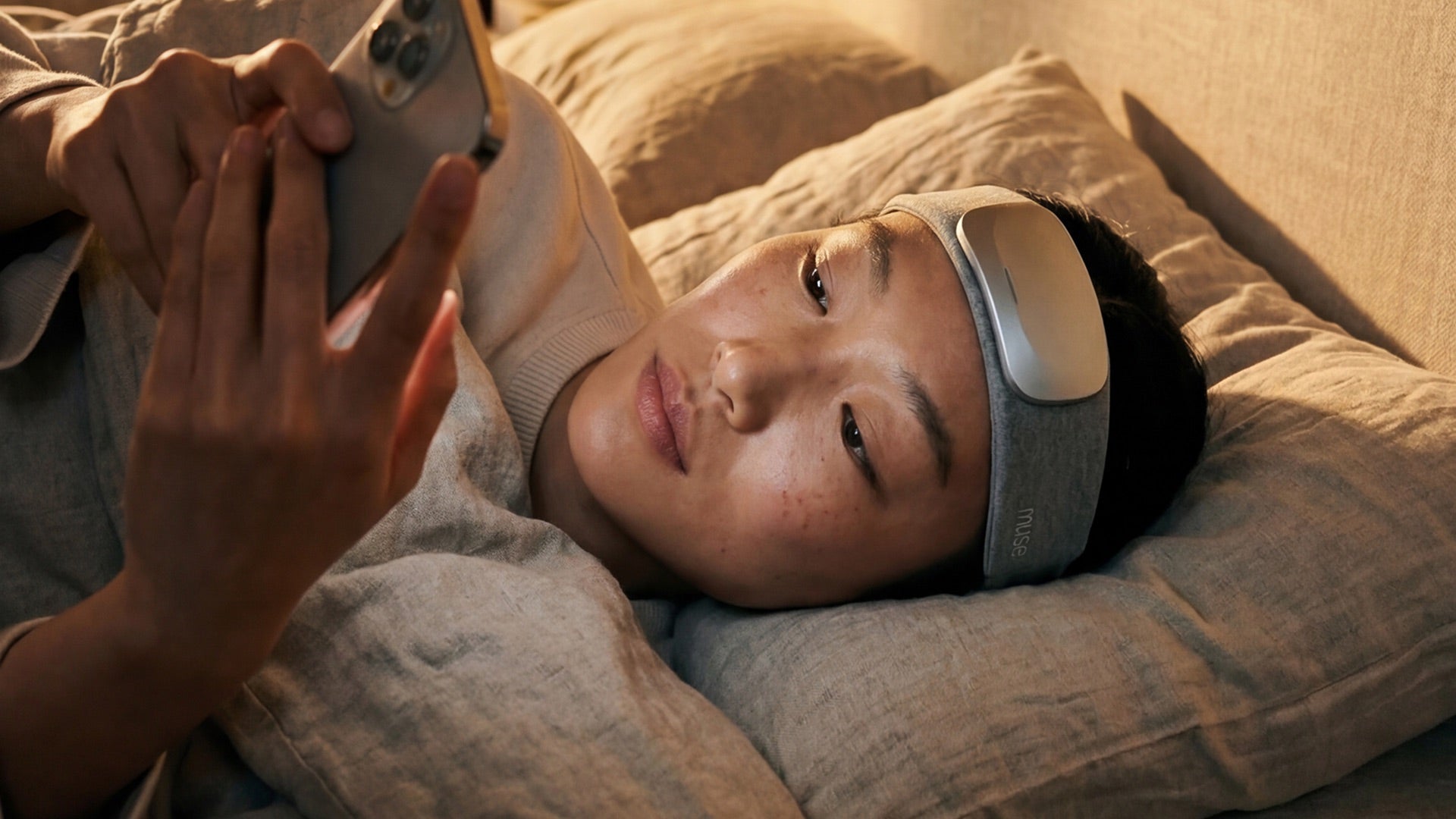 A woman lying in bed wearing the Muse S Athena soft headband, checking her phone